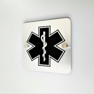 May include: A silver metal sign with a black Star of Life symbol and a snake wrapped around a staff. The sign has two silver screws in the top corners.