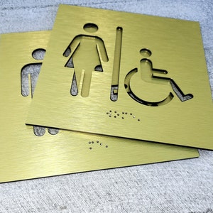 May include: Two gold metal signs with cut-out symbols for a woman, a man, and a person in a wheelchair. The signs also have braille markings.