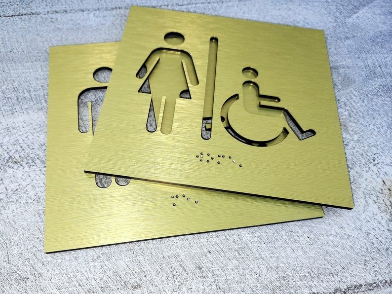 May include: Two gold metal signs with cut-out symbols for a woman, a man, and a person in a wheelchair. The signs also have braille markings.