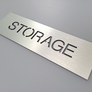 May include: A silver metal sign with the word "STORAGE" cut out in the center.