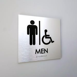 May include: A silver metal sign with black text and symbols. The sign reads "MEN" with a male stick figure and a wheelchair symbol. The sign also has braille text.