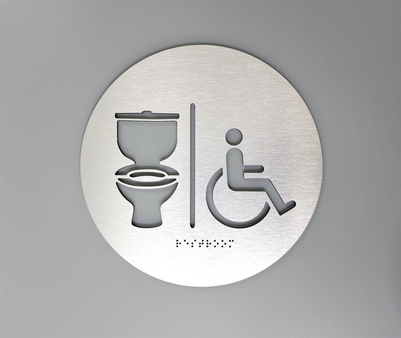 May include: A round, silver-colored sign with a toilet and wheelchair symbol. The sign also includes braille text.