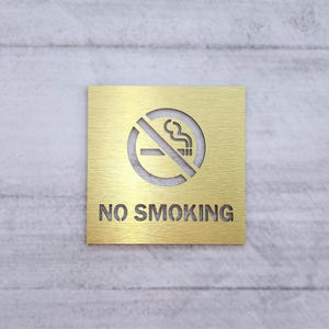 May include: A gold metal sign with a no smoking symbol and the text "NO SMOKING" on a white textured wall.