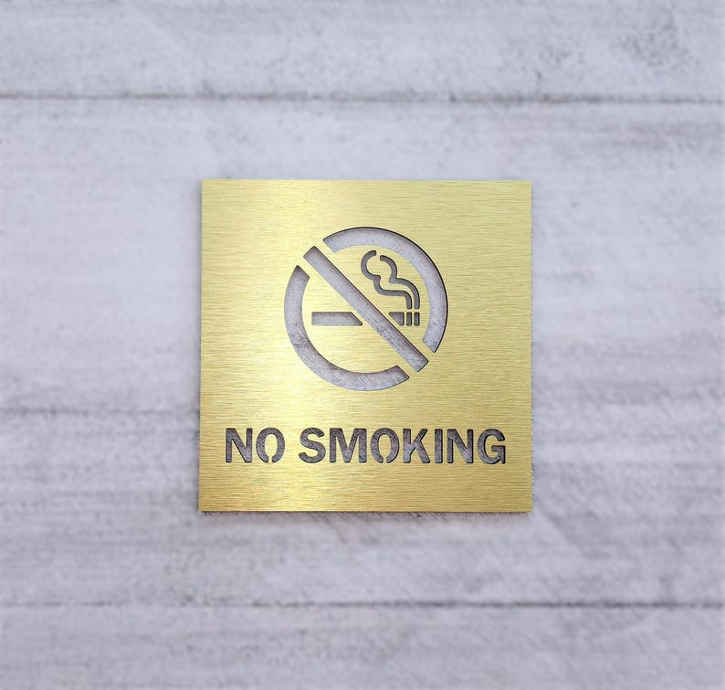 May include: A gold metal sign with a no smoking symbol and the text "NO SMOKING" on a white textured wall.