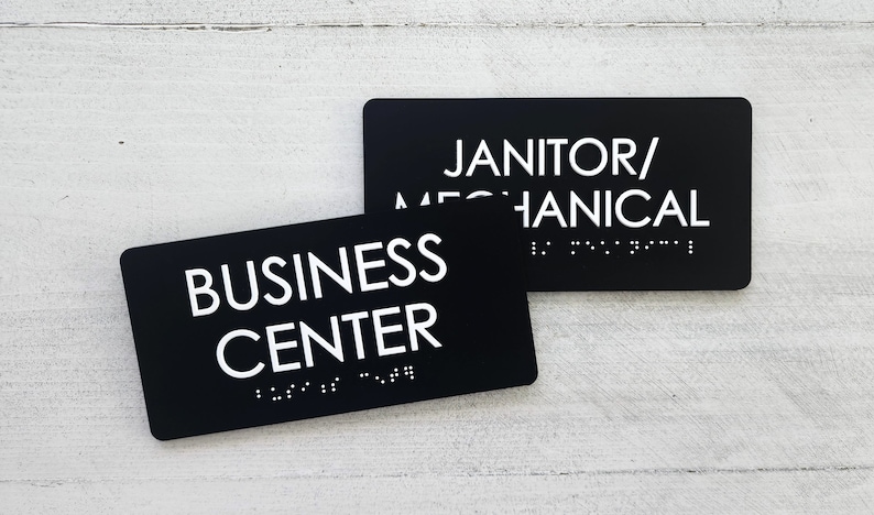 K&ouml;nnte beinhalten: Zwei schwarze rechteckige Schilder mit wei&szlig;em Text und Braille. Das obere Schild lautet "Janitor/Mechanical". Das untere Schild lautet "Business Center".