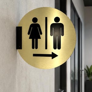 Puede incluir: Un letrero circular de metal dorado con siluetas negras de una persona con vestido y otra con pantalones, separadas por una línea vertical. Una flecha negra que apunta a la derecha se sitúa bajo las figuras, montada con un soporte de color negro.