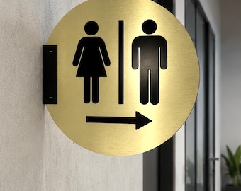 May include: A circular gold-toned metal sign featuring black silhouettes of a person in a dress and a person in trousers, separated by a vertical line. A black arrow pointing to the right is positioned below the figures, and a black bracket mounts it.