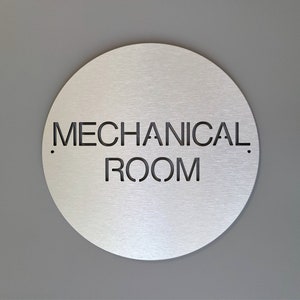 May include: A circular, brushed metal sign displaying the words "MECHANICAL ROOM" in black cut-out lettering. The sign is mounted on a grey wall. A minimalist design, suitable for industrial or commercial use.