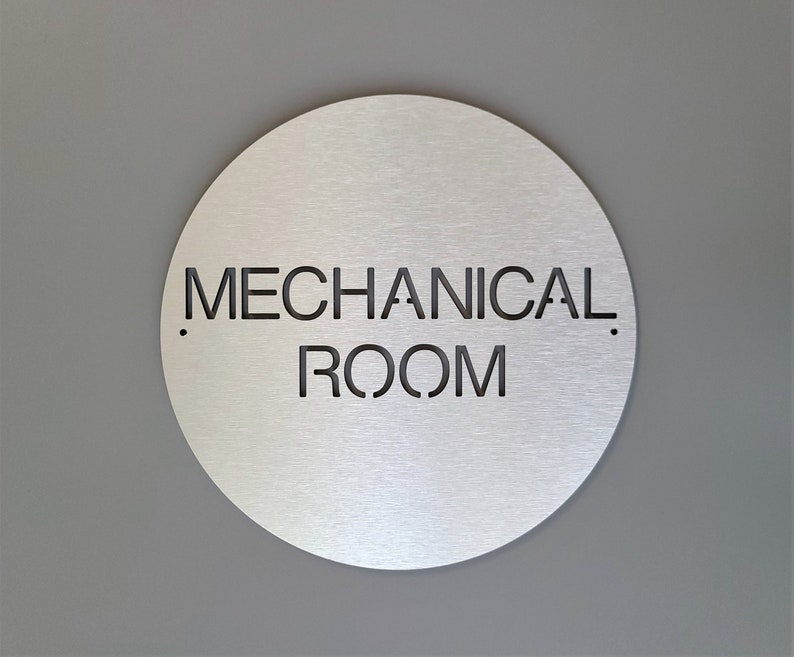 May include: A circular, brushed metal sign displaying the words "MECHANICAL ROOM" in black cut-out lettering. The sign is mounted on a grey wall. A minimalist design, suitable for industrial or commercial use.