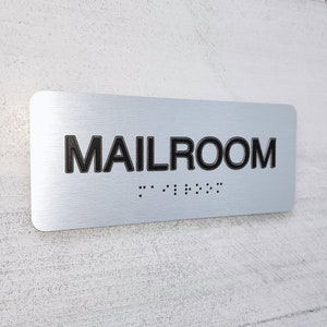 K&ouml;nnte beinhalten: Ein silbernes Metallschild mit der Aufschrift "MAILROOM" in schwarzen Buchstaben und Braille-Punkten darunter.