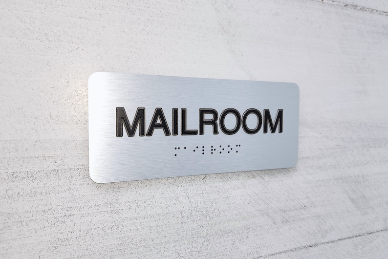 K&ouml;nnte beinhalten: Ein silbernes Metallschild mit der Aufschrift "MAILROOM" in schwarzen Buchstaben und Braille-Punkten darunter.