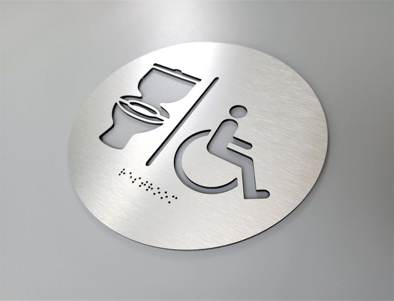 May include: A round, silver-colored metal sign with a toilet and wheelchair symbol. The sign also includes braille text.