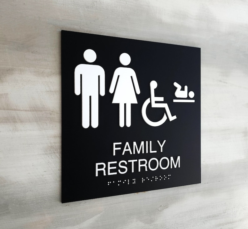 Pu&ograve; includere: Un cartello bianco e nero con il testo "FAMILY RESTROOM" in lettere bianche. Il cartello include anche icone per un uomo, una donna, una persona su sedia a rotelle e un bambino. Il cartello ha lettere in braille sotto il testo.