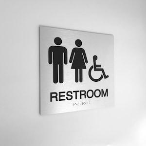 May include: A brushed metal restroom sign with black pictograms for male, female, and wheelchair accessible. The word "RESTROOM" is printed in black, with braille dots below.