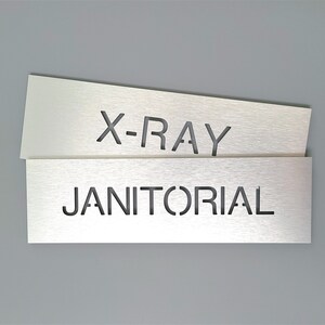 May include: Two silver metal signs with cut-out lettering. The top sign says "X-RAY" and the bottom sign says "JANITORIAL".