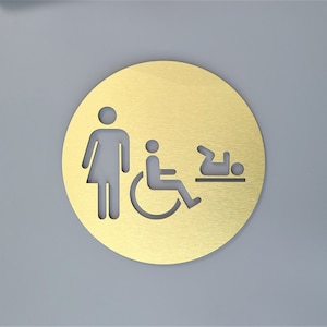 May include: A gold round sign with a white silhouette of a person, a person in a wheelchair, and a baby lying on its back. The sign is for a restroom.