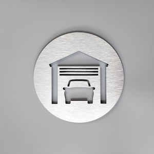 May include: A silver metal sign with a cut-out design of a garage door with a car inside. The garage door has a horizontal striped pattern.