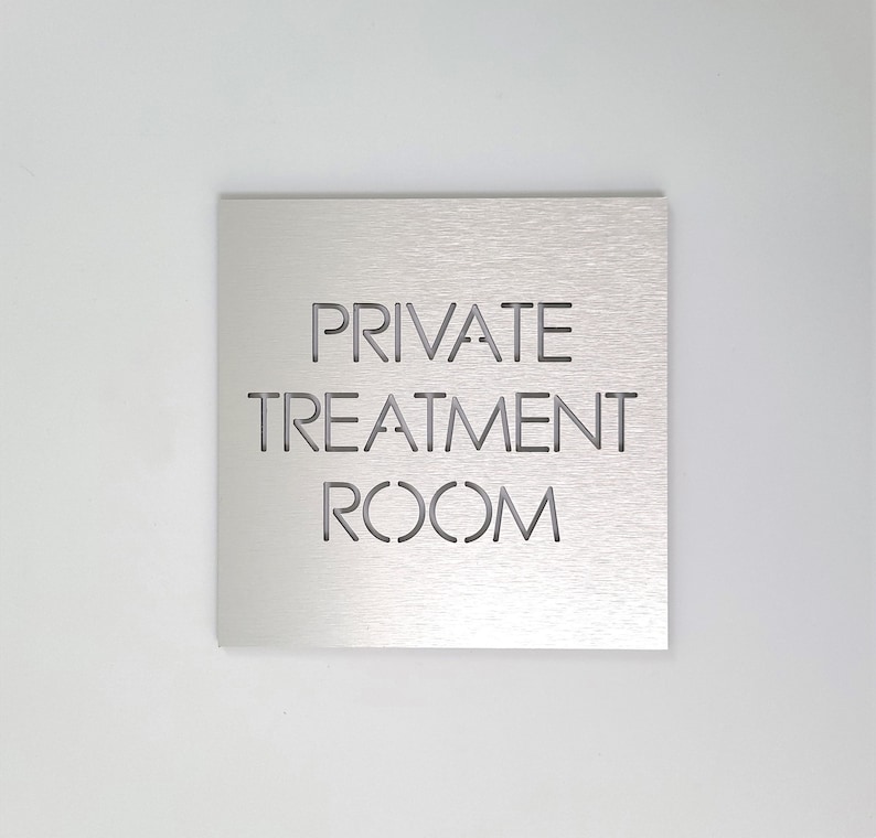 May include: A square, brushed metal sign with the words "PRIVATE TREATMENT ROOM" cut out in a sans-serif font. The sign is centered against a plain white background, creating a clean and modern aesthetic.