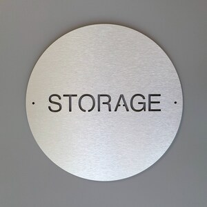 May include: A circular, brushed metal sign displaying the word "STORAGE" in black, cut out from the surface. Small black dots flank the word. The sign is mounted on a grey wall.