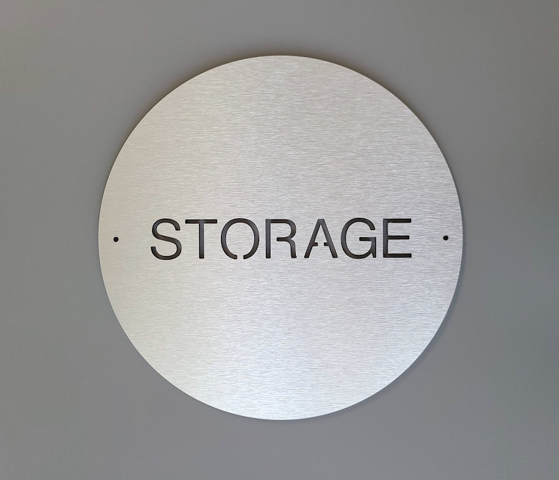 May include: A circular, brushed metal sign displaying the word "STORAGE" in black, cut out from the surface. Small black dots flank the word. The sign is mounted on a grey wall.