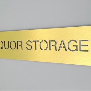 May include: A gold metal sign with the words "LIQUOR STORAGE" cut out.