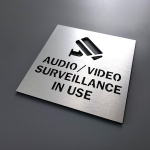May include: Silver metal sign with black text that reads "Audio/Video Surveillance In Use". A black silhouette of a security camera is cut out of the sign.