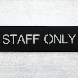 Op de afbeelding: Zwart rechthoekig bord met de tekst "STAFF ONLY" in witte uitgesneden letters.