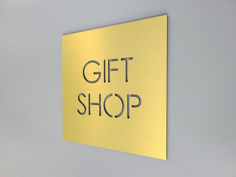 May include: A gold-colored rectangular sign with the words "GIFT SHOP" cut out. The sign is mounted on a light gray wall. The sign has a brushed metal texture.