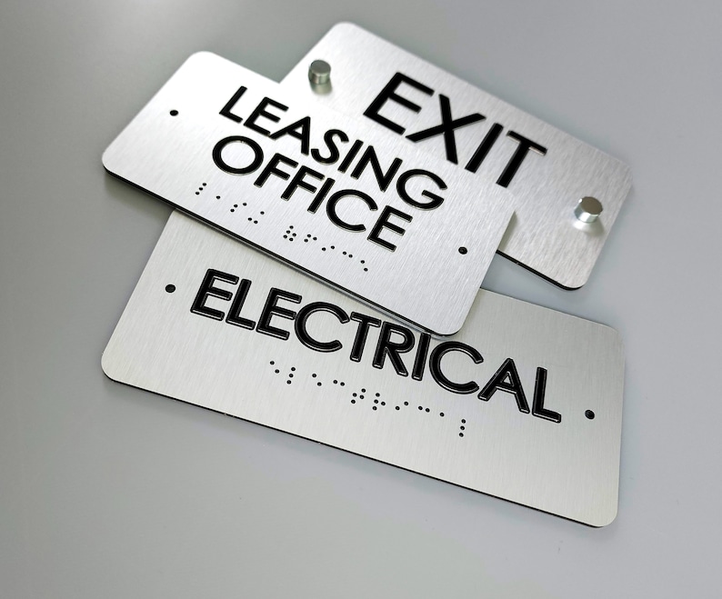 K&ouml;nnte beinhalten: Zwei silberne Metallschildchen mit schwarzem Text und Braille-Punkten. Das obere Schild zeigt "EXIT LEASING OFFICE" und das untere Schild zeigt "ELECTRICAL".