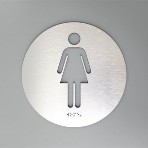 May include: A round, silver sign with a cutout of a female figure. The sign has braille lettering at the bottom.