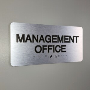 K&ouml;nnte beinhalten: Silbernes, geb&uuml;rstetes Metallschild mit schwarzem Text, der "MANAGEMENT OFFICE" lautet, und Braille-Schrift darunter.