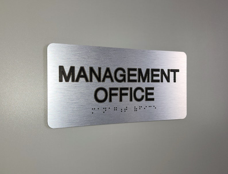 K&ouml;nnte beinhalten: Silbernes, geb&uuml;rstetes Metallschild mit schwarzem Text, der "MANAGEMENT OFFICE" lautet, und Braille-Schrift darunter.