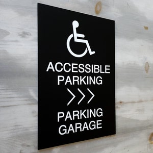 May include: Black and white sign with the words "Accessible Parking" and "Parking Garage" with a wheelchair symbol and three arrows pointing to the right.