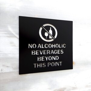 May include: Black metal sign with a white silhouette of a crossed out wine glass and bottle. The sign reads "NO ALCOHOLIC BEVERAGES BEYOND THIS POINT."