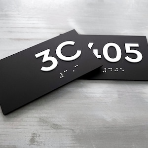 May include: Black rectangular sign with white raised numbers and braille lettering reading "3C 05".