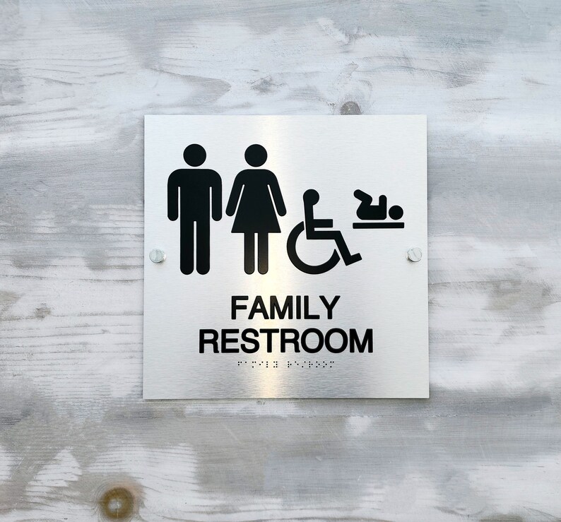 Pu&ograve; includere: Un cartello bianco con testo nero e simboli che recita "Family Restroom". Il cartello include icone per un uomo, una donna, una persona su sedia a rotelle e un bambino.