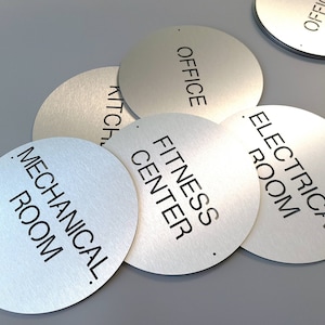 May include: Brushed metal circular signs with black text, including "OFFICE", "KITCHEN", "MECHANICAL ROOM", "FITNESS CENTER", and "ELECTRICAL ROOM". Each sign has a small hole. Ideal for commercial or office spaces.