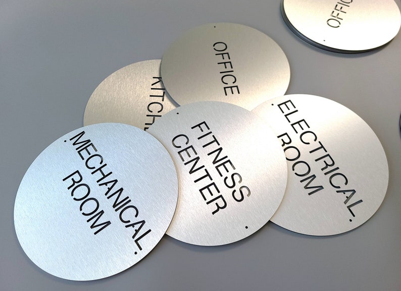 May include: Brushed metal circular signs with black text, including "OFFICE", "KITCHEN", "MECHANICAL ROOM", "FITNESS CENTER", and "ELECTRICAL ROOM". Each sign has a small hole. Ideal for commercial or office spaces.