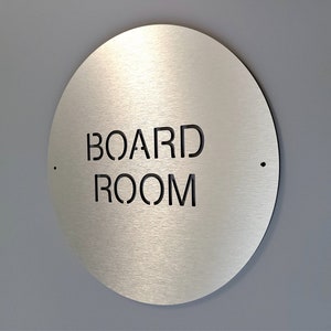 May include: A circular, brushed metal sign with the words "BOARD ROOM" cut out in black, mounted on a grey wall. The sign has a minimalist design with two small holes on either side. The sign is a decorative element for an office.