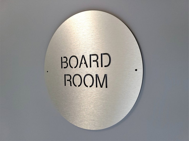 May include: A circular, brushed metal sign with the words "BOARD ROOM" cut out in black, mounted on a grey wall. The sign has a minimalist design with two small holes on either side. The sign is a decorative element for an office.