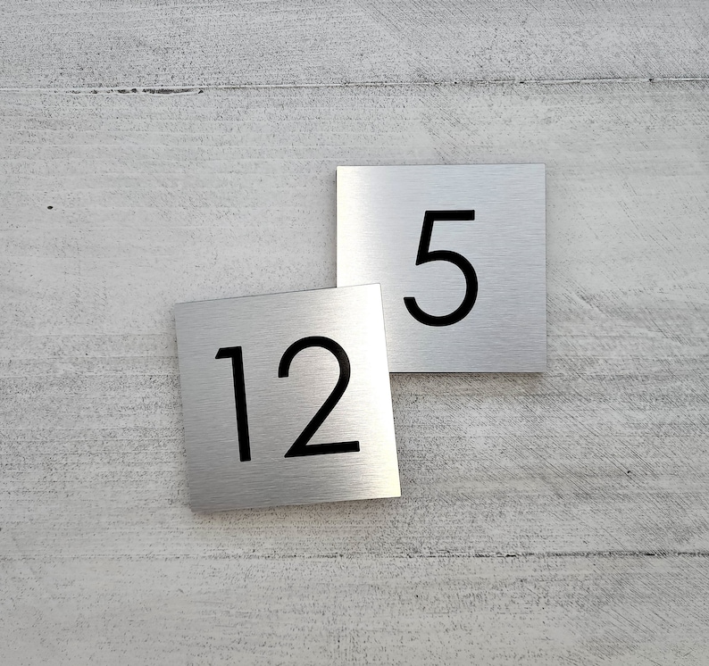 May include: Two silver metal house number plaques with black numbers. The top plaque has the number "5" and the bottom plaque has the number "12".
