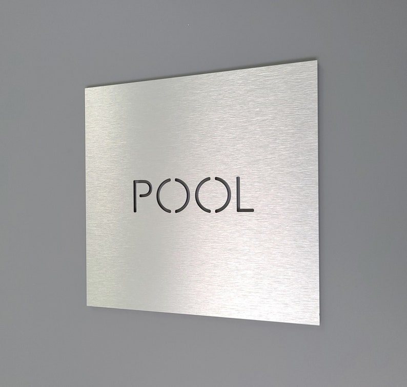 May include: A square, brushed metal sign with the word "POOL" cut out in a sans-serif font. The sign is mounted on a gray wall. The sign is a decorative element for a pool area.