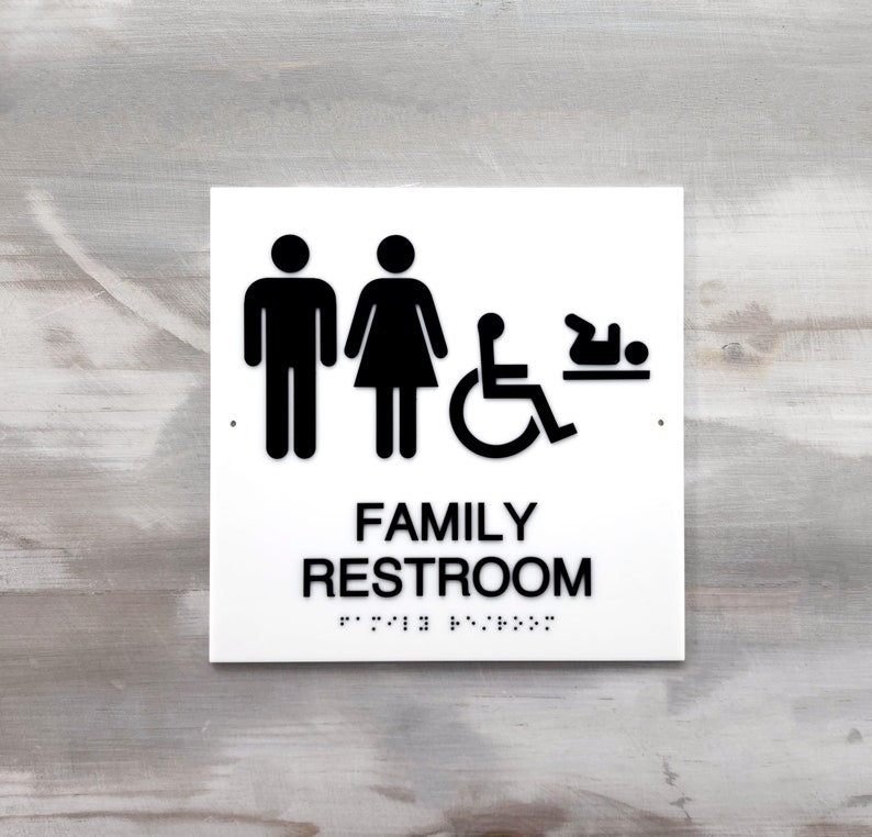 Pu&ograve; includere: Un cartello bianco con simboli neri per un bagno per famiglie. I simboli includono un uomo, una donna, una sedia a rotelle e un bambino. Il cartello dice anche "FAMILY RESTROOM" in testo nero.