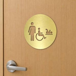May include: A circular gold-toned metal sign featuring cut-out icons of a standing figure, a person in a wheelchair, and a baby on a changing table. The sign is mounted on a light-colored wooden door with a silver-toned metal door handle below it.