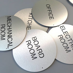 May include: A selection of circular brushed metal signs with black lettering. The signs are labelled "OFFICE", "KITCHEN", "MECHANICAL ROOM", "BOARD ROOM", and "ELECTRIC ROOM". The signs are overlapping each other.