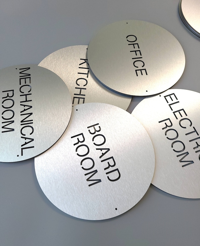 May include: A selection of circular brushed metal signs with black lettering. The signs are labelled "OFFICE", "KITCHEN", "MECHANICAL ROOM", "BOARD ROOM", and "ELECTRIC ROOM". The signs are overlapping each other.