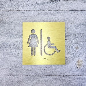 May include: A gold metal sign with a woman and wheelchair accessibility symbol. The sign also includes braille lettering.