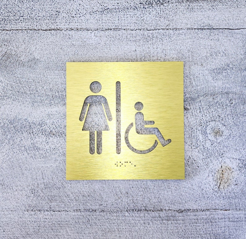 May include: A gold metal sign with a woman and wheelchair accessibility symbol. The sign also includes braille lettering.