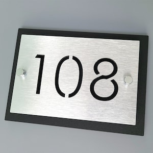 May include: A black framed silver house number plaque with the number 108 cut out in black. The plaque has two silver screws on the sides.