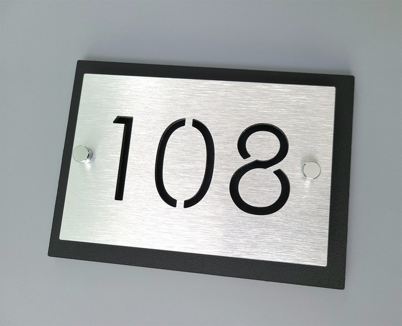 May include: A black framed silver house number plaque with the number 108 cut out in black. The plaque has two silver screws on the sides.